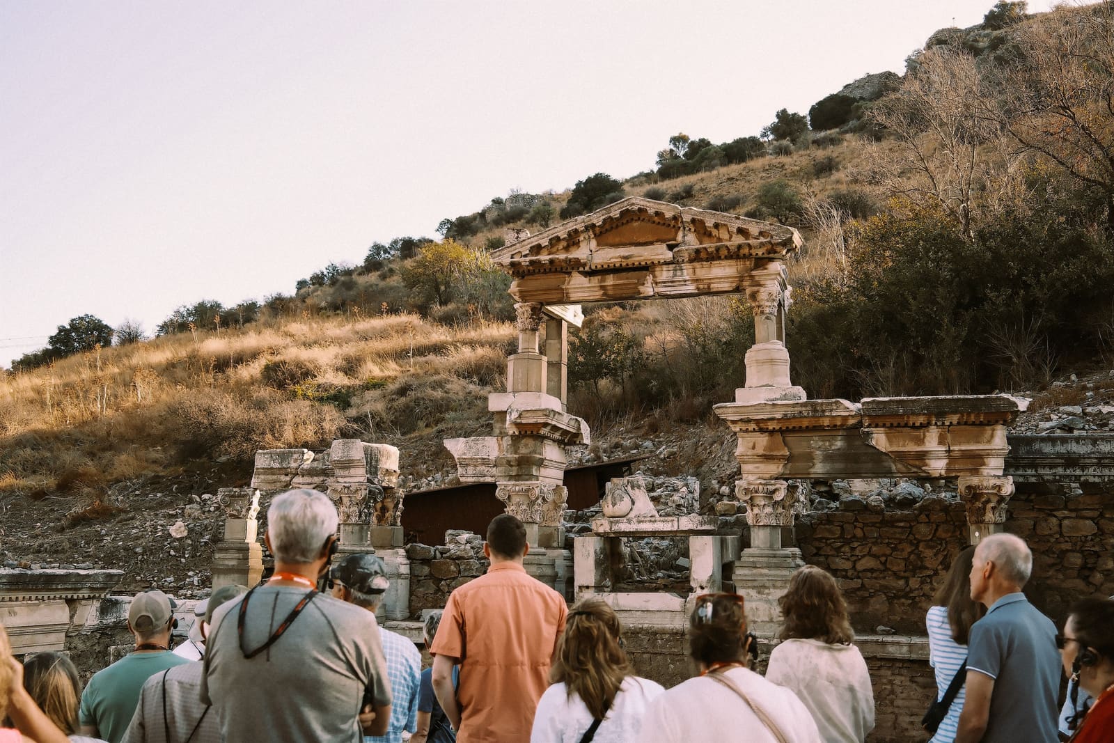 A guided group of travelers seen from behind, listening in front of the Fountain of Trajan on Curetes Street at Ephesus