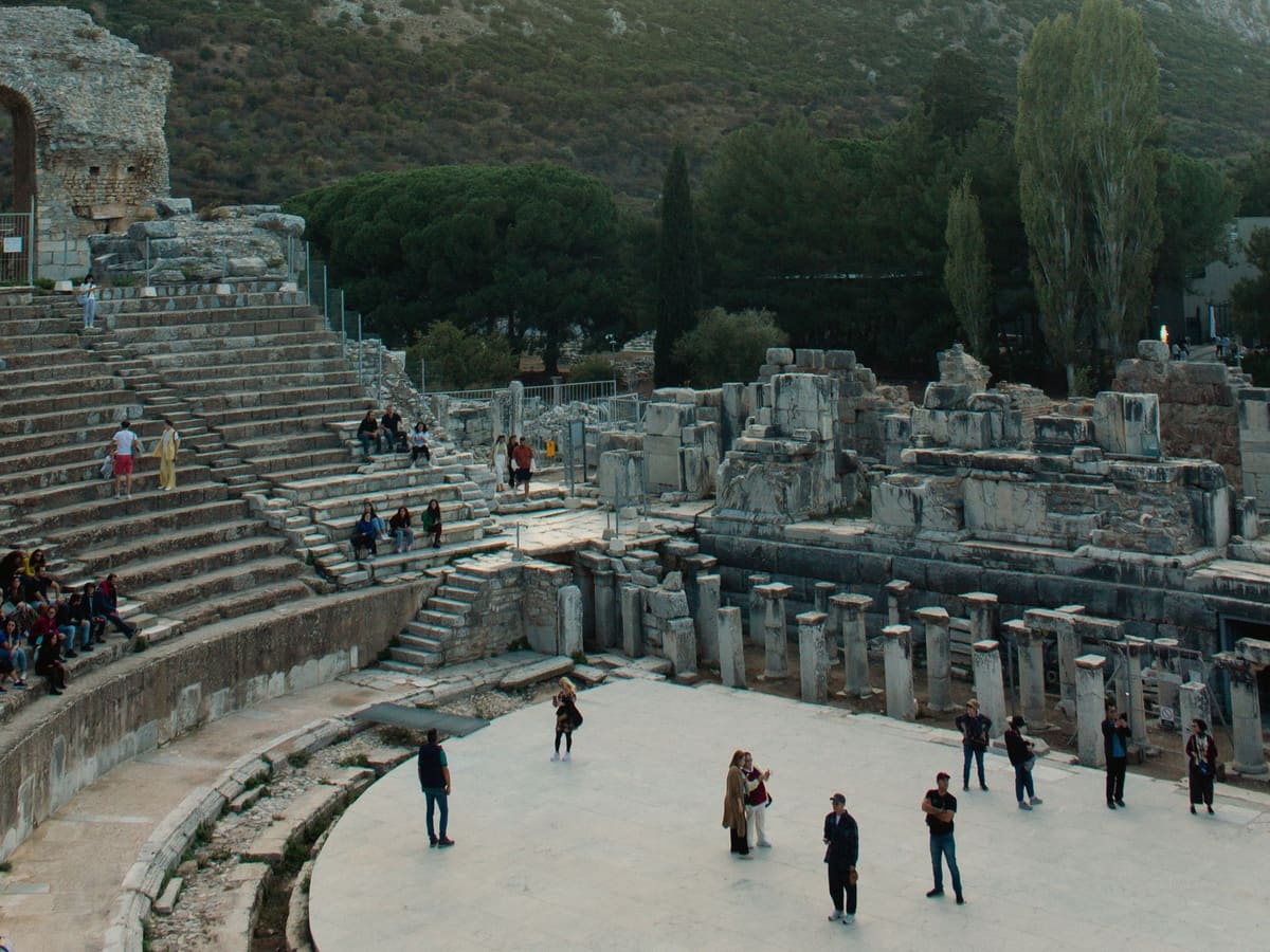 Visitors standing on the paved orchestra floor of the Great Theater of Ephesus, seen from the seating, with the stage building foundations behind