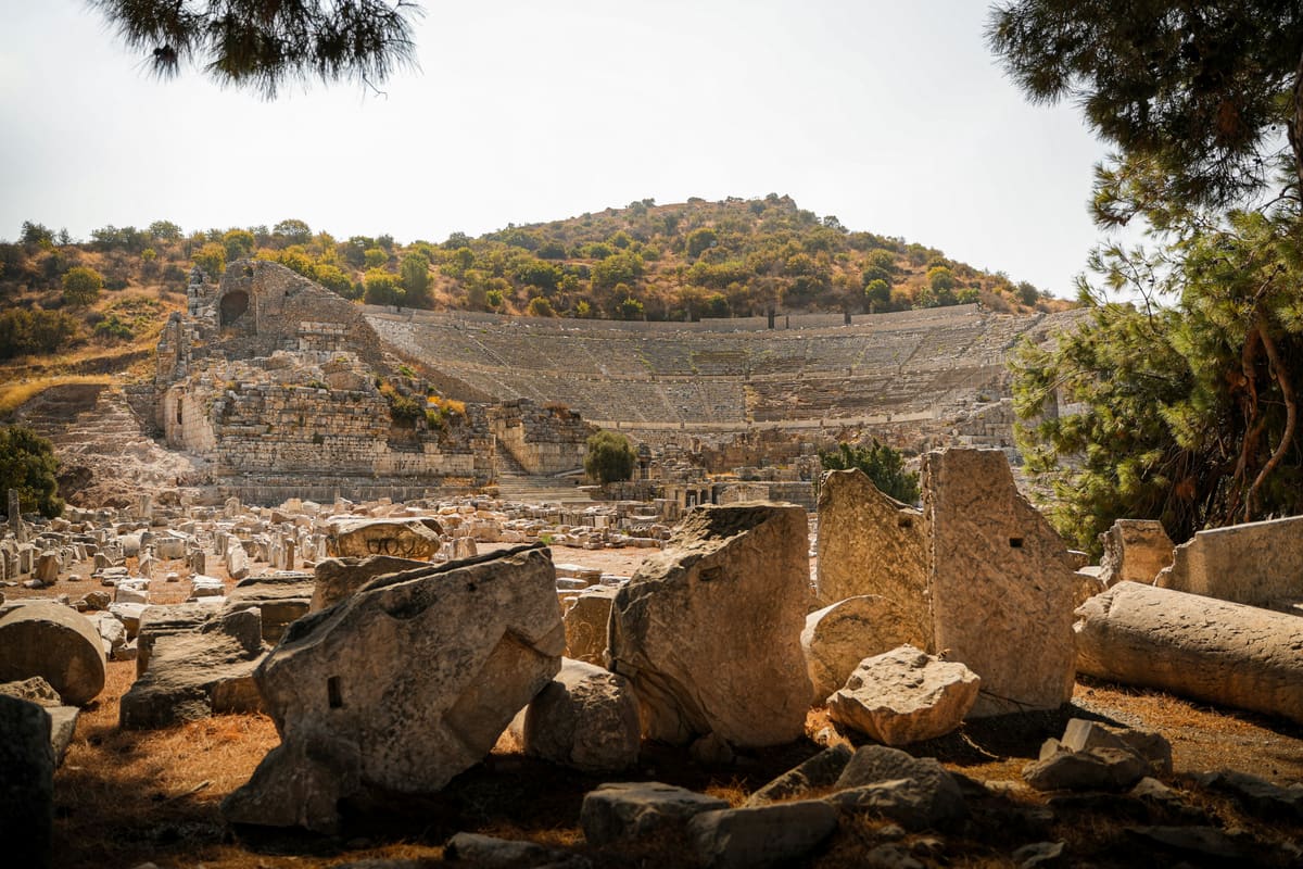 The Great Theater of Ephesus seen across fallen marble blocks, its curved tiers of stone seats rising up the hillside behind the ruined stage building