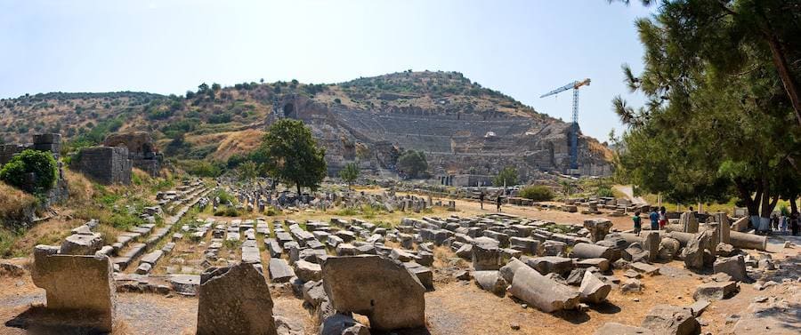 The Great Theater of Ephesus on its hillside above fallen stone blocks in the lower city, with a construction crane beside the stage building