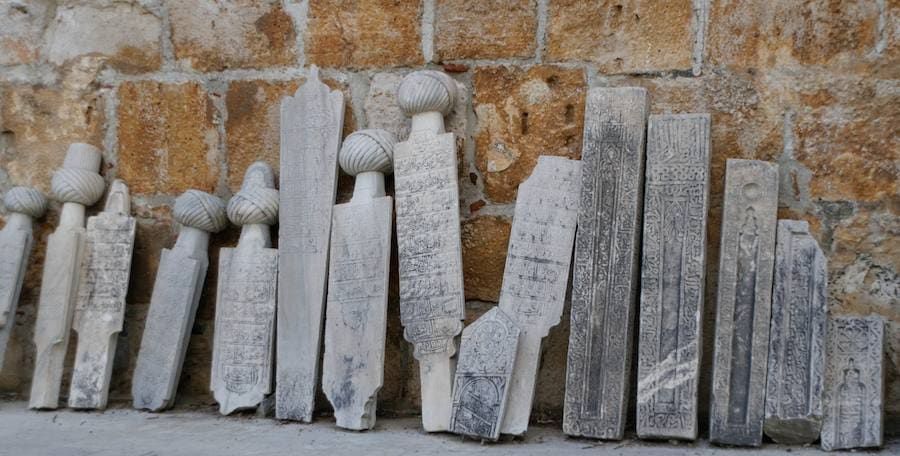 Carved Ottoman marble tombstones, several topped with stone turbans, leaning in a row against an old stone wall