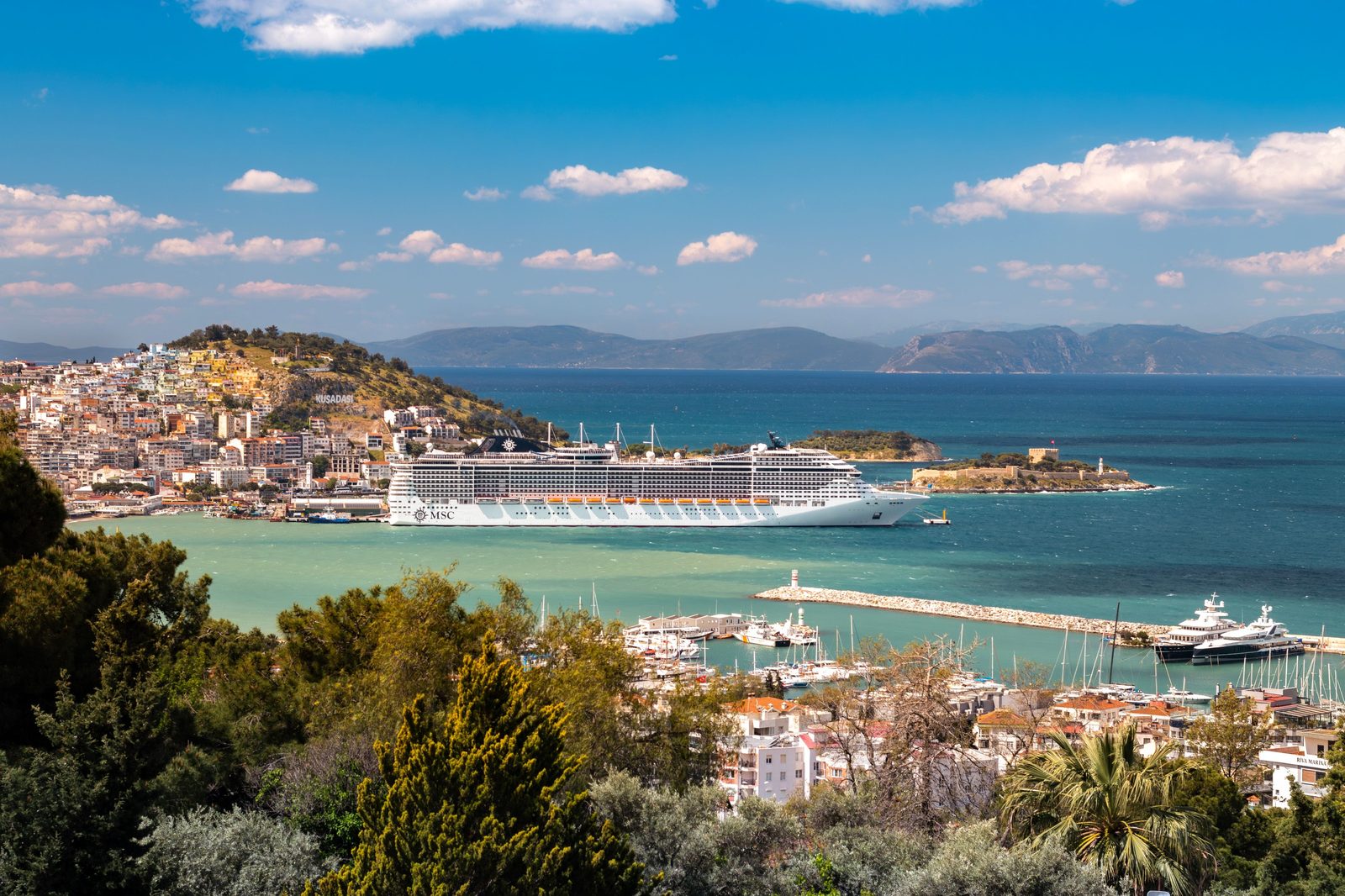 Kusadasi harbor seen from the hillside, a large cruise ship at the pier, Pigeon Island and its fortress beside it and mountains across the gulf