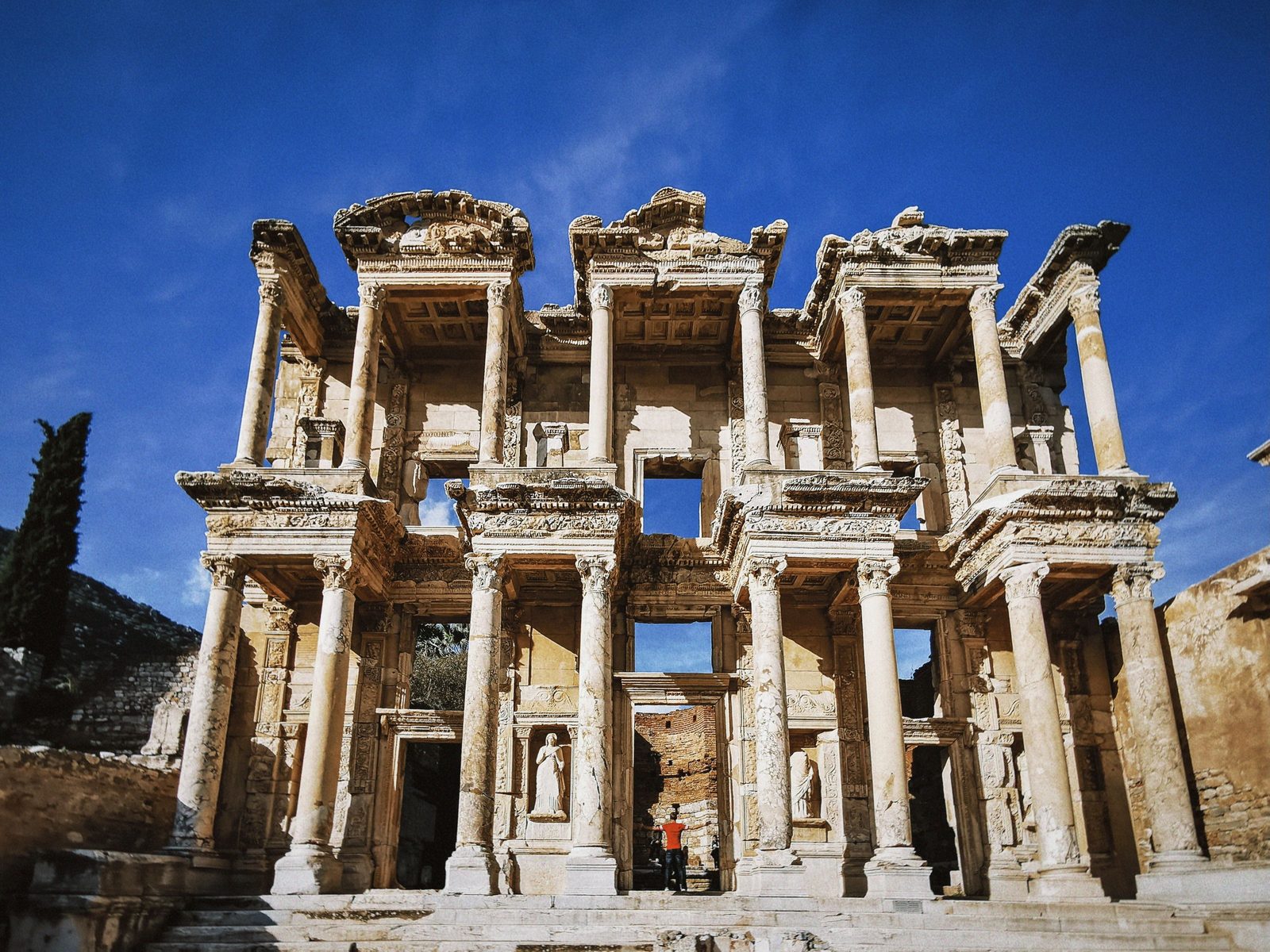 The two-story marble facade of the Library of Celsus in Ephesus against a deep blue sky, with statues in its niches and a cypress tree at the left