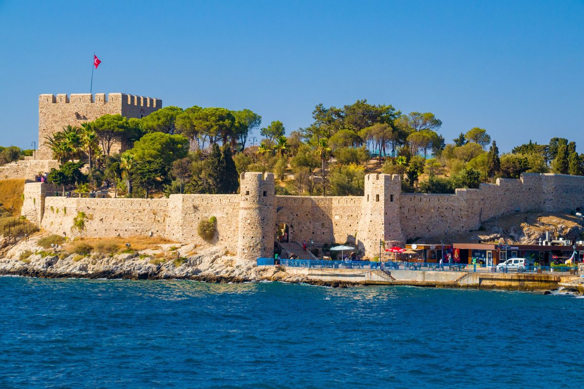 The stone walls, round towers and crenellated keep of the fortress on Pigeon Island in Kusadasi, seen from the sea with a Turkish flag on top