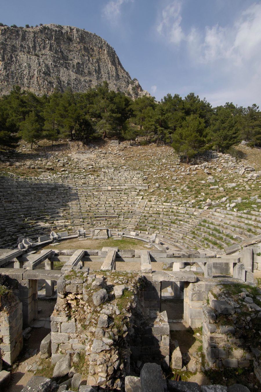The stone seating of the ancient theater of Priene rising toward pine trees, with the gray limestone cliff of Mount Mycale above