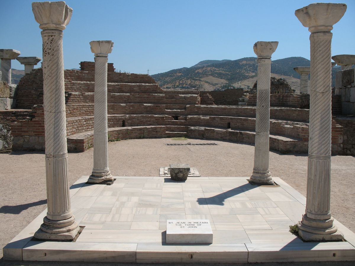 Four marble columns with spiral fluting standing at the corners of the tomb platform of Saint John, with a small marker stone in front and brick walls behind